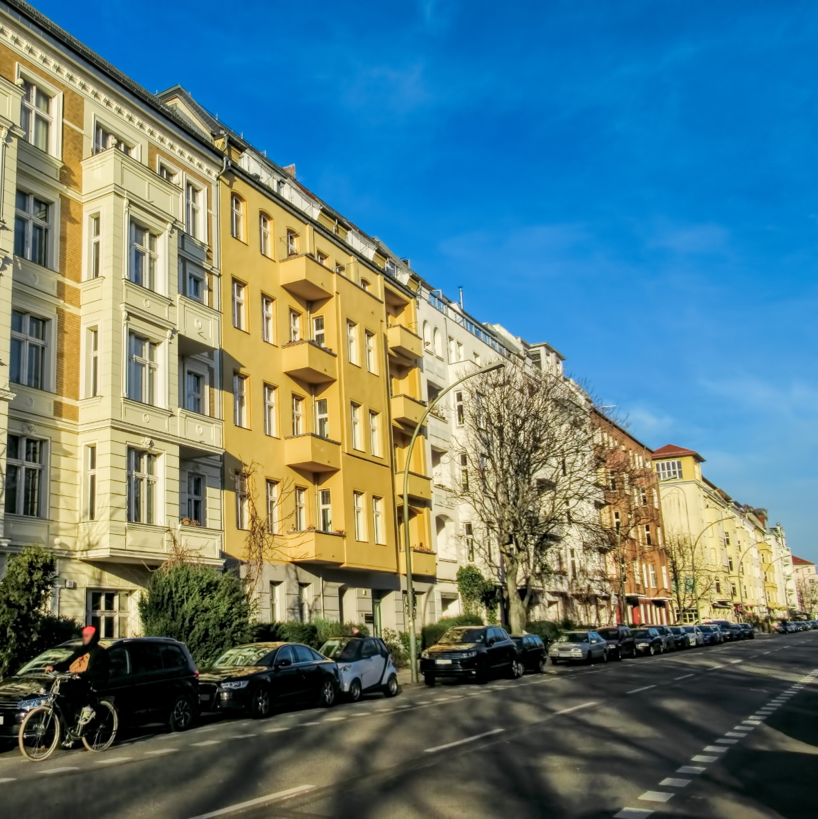 Berliner Altbauhäuser in einer Reihe, alle in verschiedenen Gelben Pastelltönen. Im Vordergrund eine Straße und ein Fahrradfahrer.