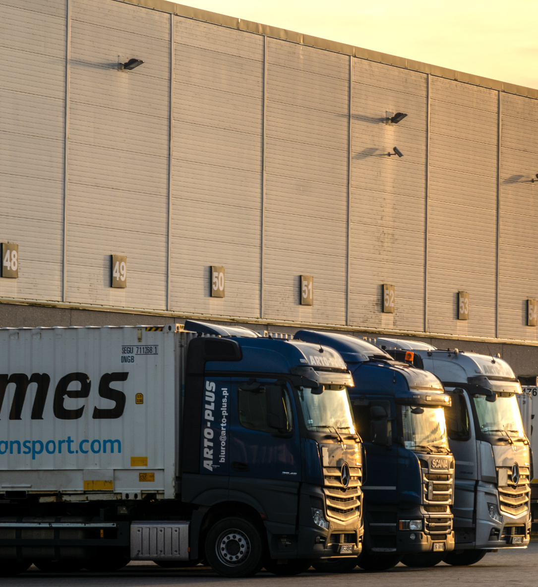 Im Vordergrund Scania und Mercedes Lastwagen, beladen mit Hermes und DHL Containern. Im Hintergrund eine Ladestation im Sonnenuntergang.