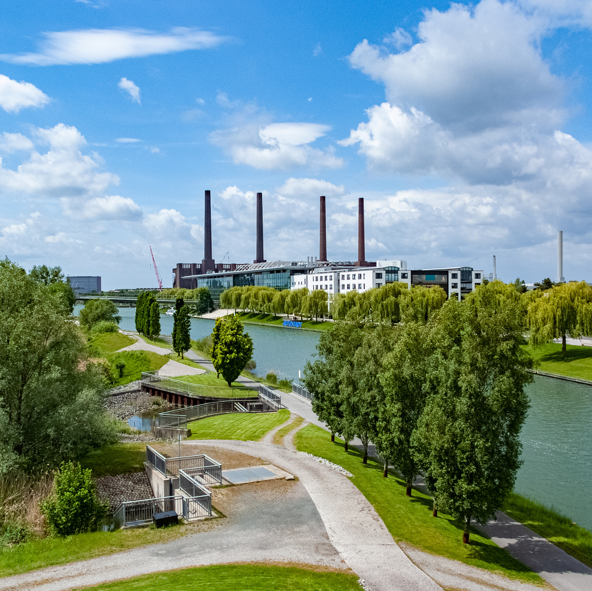 Fabriken mit vier Abdampftürmen, in wessen Vordergrund sich eine große Grünfläche, mit Wegen, Bäumen und ein Fluss befinden.